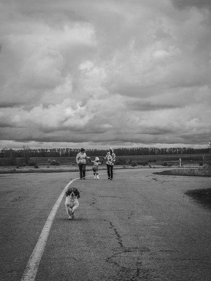 A-black-and-white-picture-of-a-family-in-the-distance-on-a-road-and-a-dog-running-head-towards-the-camera.