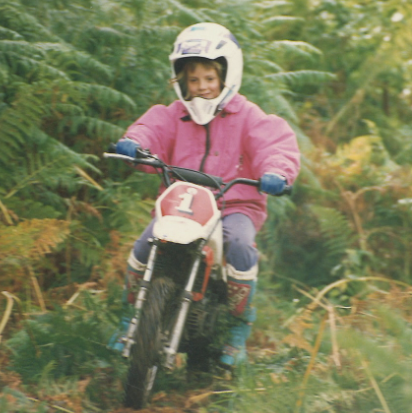 a young girl wearing a white helmet, pink jacket, jeans and coloured boots rides a motorcycle through fern bushes