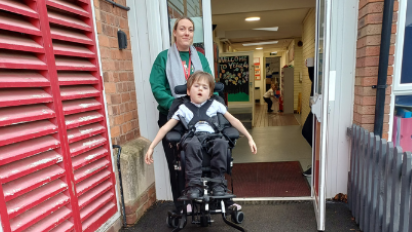 Dylan and Siani pictured in front of an open door which leads to a classroom. Siani wears a green jumper while Dylan is in a wheel chair, wears a white polo shirt and grey school trousers. 