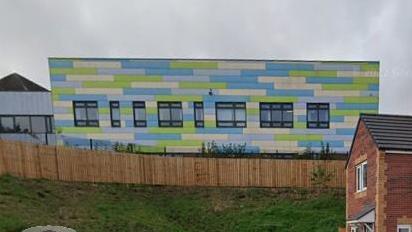 A school building with blue and green cladding seen from behind a wooden fence.