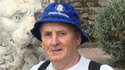 Stanley Smith smiling and wearing a blue bucket hat with the words Sergio Tacchini written on in white lettering. He appears as though he is on holiday in a park. A stone lion is behind him. He has a small stud earring and wears a white T-shirt. The straps of a black backpack can also be seen.