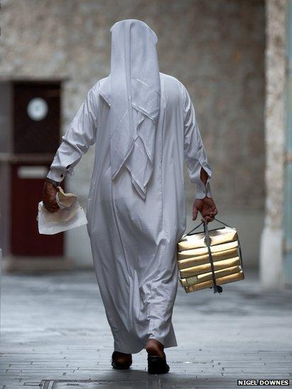 A Qatari man in traditional dress carries his lunch boxes in one hand and holds his Arabic flat bread in the other.