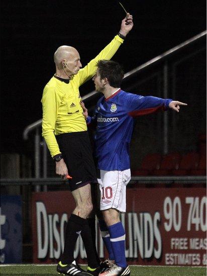 Michael Carvill receives a yellow card for diving from referee Colin Burns following the penalty claim