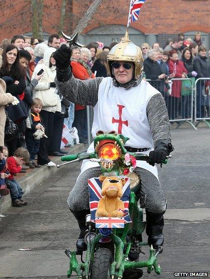 Man dressed in a St George costume riding on a scooter decorated as a dragon and displaying a bull dog toy