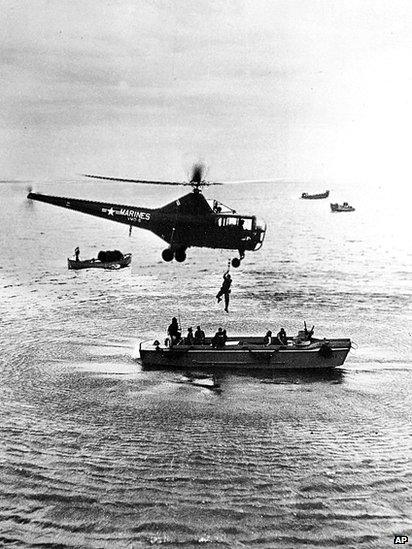 A hovering US Marine helicopter picks up personnel from a landing barge in the harbour at Inchon