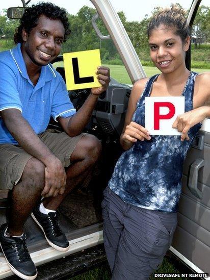 Two young Aboriginal people holding up "L" and "P" plates next to car