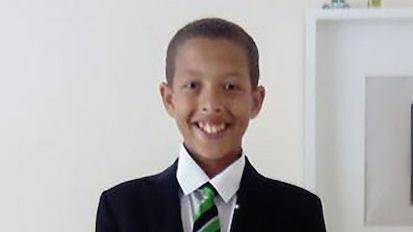 Noah smiling at the camera as he stands against a white wall. He is wearing his school uniform which is a black blazer, green and black striped tie and a white shirt. He has short black hair.