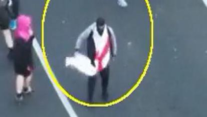 View from above of a man in a black balaclava and dark clothes with a white flag with a large red cross on it draped around his neck