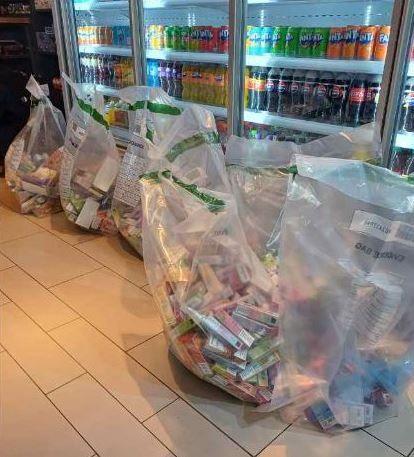 Six large see-through bags full of disposable vapes are sat on the floor of a shop which has drinks fridges behind them. 