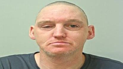 A police mugshot of a man with closely cropped hair and some grey facial hair. He is wearing a grey T-shirt.