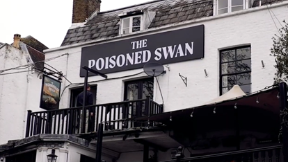 A view from street level of the pub signage. It is a large black and white wall-mounted sign saying 'The Poisoned Swan' in capital letters, on the white stone washed pub building, which has black framed windows and a black painted balcony.