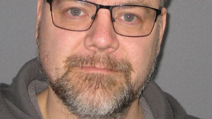 A police photo of a man with short brown hair, a moustache and a beard. He is wearing a grey hoodie and black-rimmed glasses. 