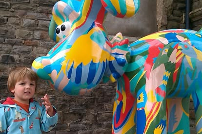 Three-year-old Barney reaches up toward a Gromit sculpture. He wears a red T-shirt under a bright blue raincoat with multicoloured details. Gromit is painted in a mix of vivid colours and patterns.
