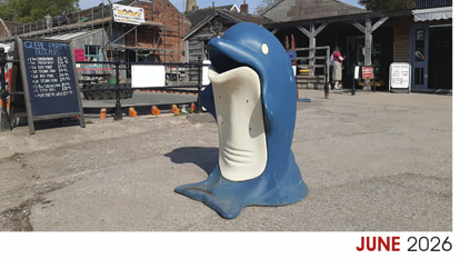 A blue and white bin, shaped like a dolphin. Behind it are a number of buildings, some wooden framed.