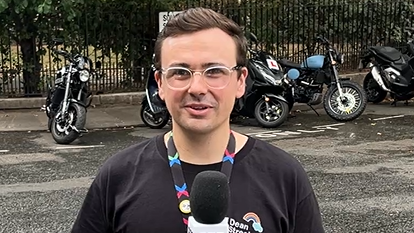 Lawrence O'Connell is seen wearing glasses and a black T-shirt is holding a microphone while standing on a street, with several parked motorbikes and a fence with greenery in the background.