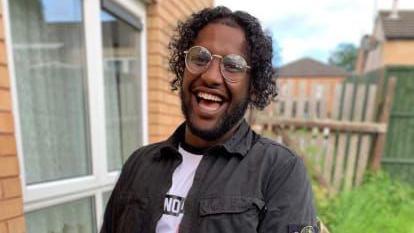 A photograph of murder victim Abdullah Hassan. He has curly black hair and a dark beard. He is wearing glasses and a dark shirt and white t-shirt. He is standing outside by a wooden fence and laughing.