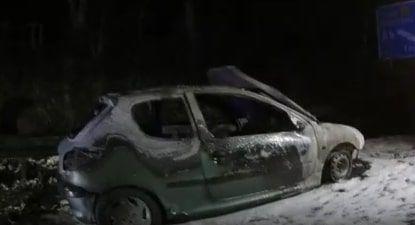 The burnt out car sitting on the side of the road. It is charred and grey.