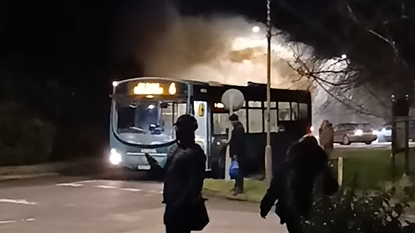 A screenshot from a video showing an evacuated bus with smoke coming out the back of it. A number of people of standing in front and to the right of the bus. There are a number of cars behind the bus and a small patch of grass to the right of the image. 