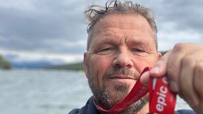 Mr Stansfield has short greying hair and beard, he is wearing a blue top and holding a medal towards the camera by a red ribbon.