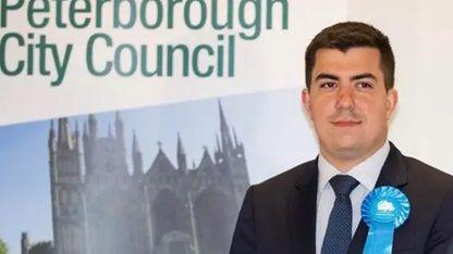 Rylan Ray stares at the camera. He wears a dark suit and a navy tie and has a Conservative party rosette. In the background is a Peterborough City Council sign with an image of the city's cathedral.