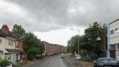 Part of Broad Lanes, which has houses and other buildings on one side and an industrial building, trees and parked cars on the other side.