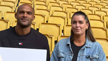 A man with a shaved head wearing a black sweater sits next to a woman wearing a light blue denim jacket in a football stadium. Behind them are rows of yellow seats