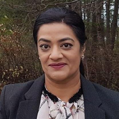 A woman with tied-up brown hair, wearing a black jacket and a pink top, looks directly at the camera. Trees can be seen in the background.