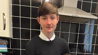Jacob, 14, with brown hair and wearing a white shirt and black crew neck jumper is photographed in a kitchen with black kitchen tiles on the wall behind him and an oven hob and kitchen cupboard.