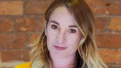 A professional headshot of a woman with long blonde hair standing in front of a brick wall. 