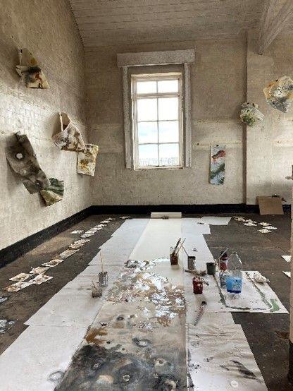 The inside of a temporary art studio on Orford Ness. On the walls are two and three-dimensional paintings. The floor is covered in white pieces of paper and paint brushes and paint pots.