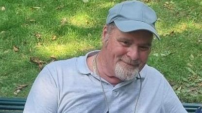 A man in a grey polo shirt and grey cap had short grey hair and facial hair. He is sat on a bench with an area of grass behind him.