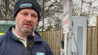 A man in a black woolly hat with the Yorkshire Water logo on it. He also wears a blue zip-up fleece and stands next to an EV charge machine.