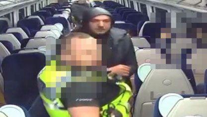 CCTV footage shows the aisle of a train where a police officer has arrested a hooded man. The faces of the officer and other passengers are pixelated.