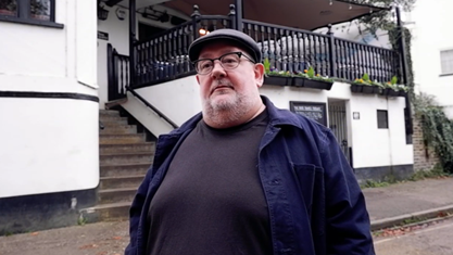 Johnny Vegas stands outside the pub wearing a dark jumper and coat, flat cap and glasses