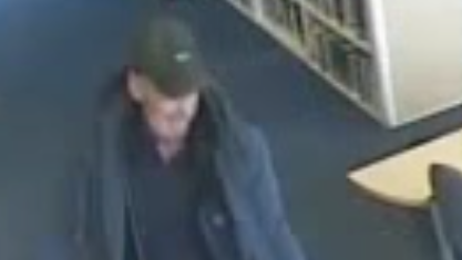A closer shot of the man, who can be seen in a dark cap, black or navy jacket and black or navy shirt. He is looking down, away from the camera. A library table and a set of shelves with books on them can be seen in the background.