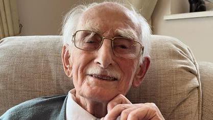 Man wearing glasses looks straight into the camera lens. He's smiling and wearing a white shirt with a blue and white spotted tie and grey cardigan