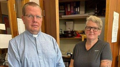 A man, wearing a dog collar and a blue shirt with a stern expression standing next to a woman wtih short blonde hair and glasses