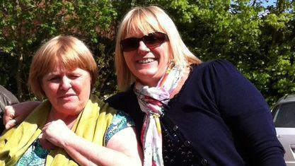 Shelagh Goodwin and Lindsay Murphy, standing side by side, looking at the camera. Shelagh, on the left is Shelagh, squinting her eyes, wearing a yellow scarf and green top, Lindsay is next to her wearing sunglasses, a multicoloured scarf and blue top and cardigan. They are standing in front of trees. 