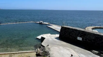 The bathing pool at La Vallette which is sheltered from the open ocean with a stone wall. There is a low diving platform at the back of the pool. The sea is calm and blue. 