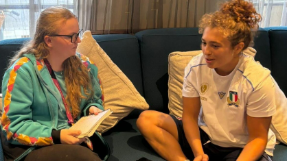 The image shows two people sitting on a blue sofa in what appears to be a lounge or waiting area. One person is wearing a bright turquoise jacket with colourful stripes on the sleeves and is holding a notepad and pen. The other person is dressed in a white sports shirt with a logo and dark shorts.