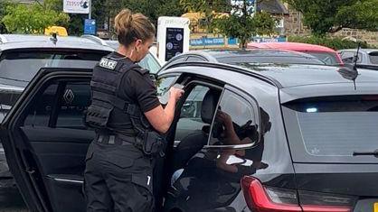 A female officer in black uniform stood by the door of a black car looking at something in her hand. 