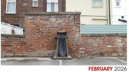 A metal framed bin, containing a black plastic bag. It is up against a brick wall at the back of terraced properties