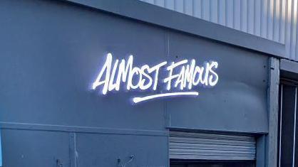 A neon sign for 'Almost Famous' on the front of a grey building above the entrance way to the eatery on Parr Street in Liverpool. 