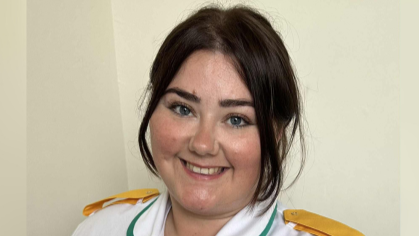 Kellie-Ann is in her white nursing uniform with yellow pads on her shoulders. She has dark brown hair which is tied back, dark eyebrows and blue eyes.