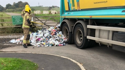 Blue refuse lorry with pile of plastic waste in a layby. There are two fire officers attending to the waste. There are trees and grass.
