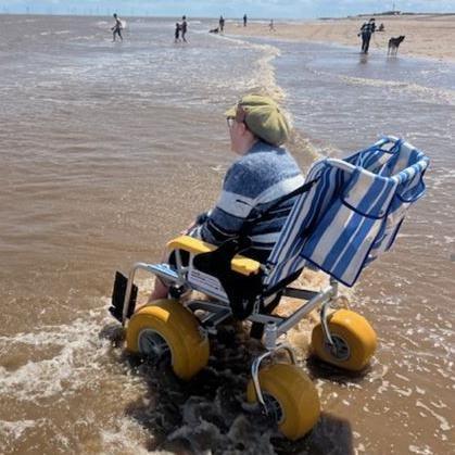 A woman is wearing glasses, a green cap and a horizontal-striped blue jumper. She is sat in a blue-and-white striped beach wheelchair, which has large yellow wheels, with her feet in the sea. She is looking out to sea.