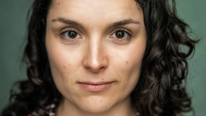 A professional headshot of Hollie. She has dark brown, curly hair and dark brown eyes. She is smiling slightly. Behind her is a sage green background.