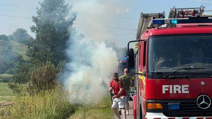 Images of two people one in shorts and t-shirt pulling a hose from the back of a fire engines as there is smoke pouring into the sky behind them in a grassy area