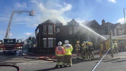 Fire crews stood outside the home which smoke was pouring out of, with a fire engine parked to the left of the picture with a firefighter hoisted above the fire on a platform and another to the right of the picture 