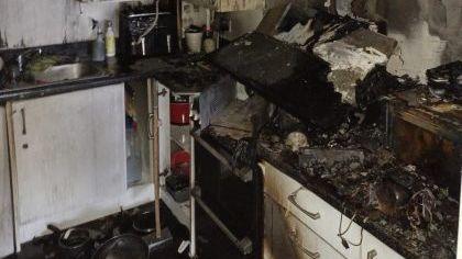 A kitchen which has melted worktops and is covered in black soot. The cooker hood has collapsed and fallen off the wall and onto the hob below. The white walls are grey with soot.